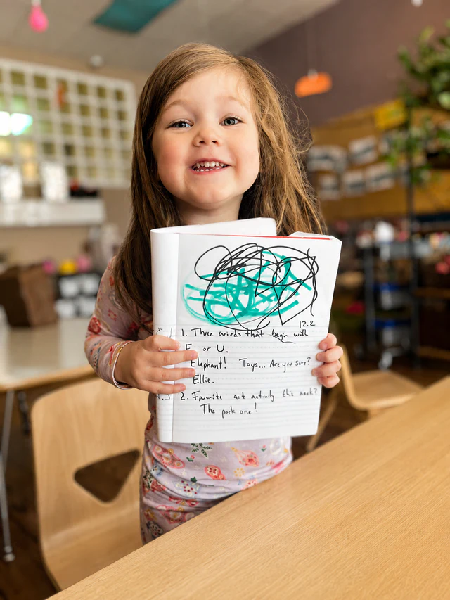 A smiling child proudly holding up her drawing in a Lakeview classroom.
